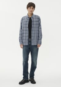 Jaunas vyras, apsirengęs mėlynos tartano rašto marškiniais su mygtukais virš juodo marškinėlio, mėlyniais džinsais ir juodais batais, stovi prieš balto fono.