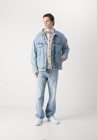 Lichtblauwe denimjack met een relaxte pasvorm, knoopsluiting aan de voorkant, twee borstzakken en bijpassende lichtblauwe jeans. Witte sneakers maken de outfit compleet.