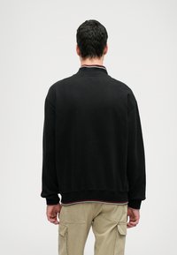 Μαύρη μπλούζα με στρογγυλή λαιμόκοψη, ριμπ μανσέτες και πολύχρωμη ριγέ τελείωμα. Το ύφασμα φαίνεται λείο με άνετη εφαρμογή.