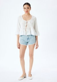 Blusa de lino blanca con escote cuadrado, frente con cordones y mangas con volantes, combinada con pantalones cortos de mezclilla azul claros y bailarinas blancas.