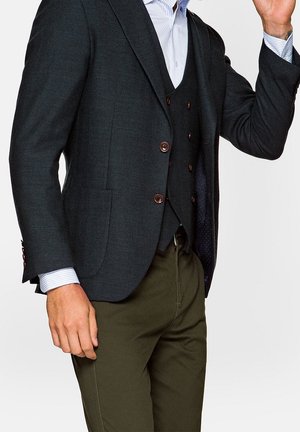 Uomo che indossa un blazer scuro e un gilet sopra una camicia a righe chiara, abbinati a pantaloni verde oliva, che alza la mano destra.