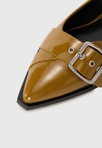 Pointed toe ankle shoe in glossy mustard yellow patent leather with a wide strap and silver buckle detail, set on a contrasting black sole.