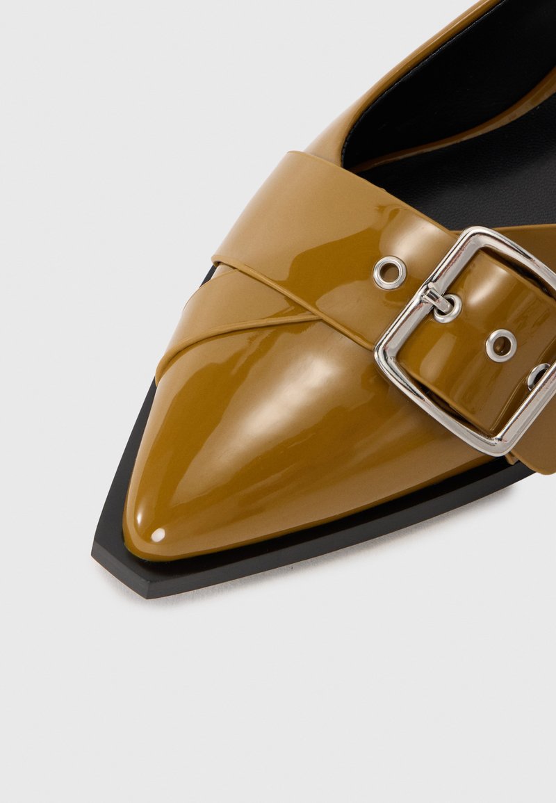 Zapato de tobillo de punta afilada en charol amarillo mostaza brillante con una amplia correa y detalle de hebilla plateada, montado sobre una suela negra contrastante.