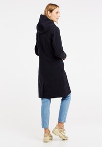 Cappotto lungo nero, impermeabile e con cappuccio. Caratterizzato da una vestibilità dritta, tasche laterali e un orlo posteriore leggermente più lungo. Indossato sopra jeans azzurri chiari e sneaker.