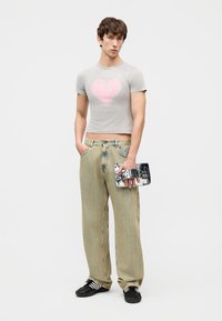 Νέος άνδρας που φοράει ένα ανοιχτό γκρι κοντό μπλουζάκι με ροζ καρδιά, μπεζ παντελόνι φαρδιάς γραμμής, μαύρα αθλητικά παπούτσια, κρατώντας μια γυαλιστερή ασημί τσάντα χειρός.