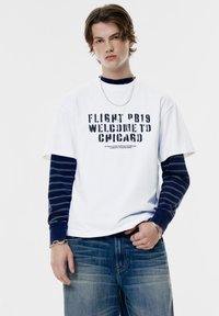 Witte t-shirt met grafische tekst "FLIGHT PB19 WELCOME TO CHICAGO," laag over een marineblauwe langwerpige shirt met een gestreept patroon. Blauwe jeans, zilveren sieraden.