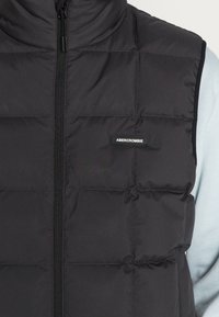Gilet imbottito nero con zip frontale, design a righe orizzontali e un'etichetta con logo sul petto. Realizzato in un tessuto liscio e opaco.