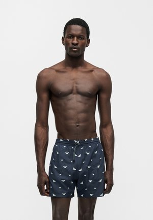 Męski model o ciemnej karnacji ubrany w granatowe kąpielówki z białymi logo orła, stojący na jednolitym jasnoszarym tle.