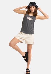 Canottiera grigia con stampa floreale, pantaloni corti crema arrotolati all'orlo e sandali a blocco neri. Cappello a secchiello in bianco e nero a scacchi.