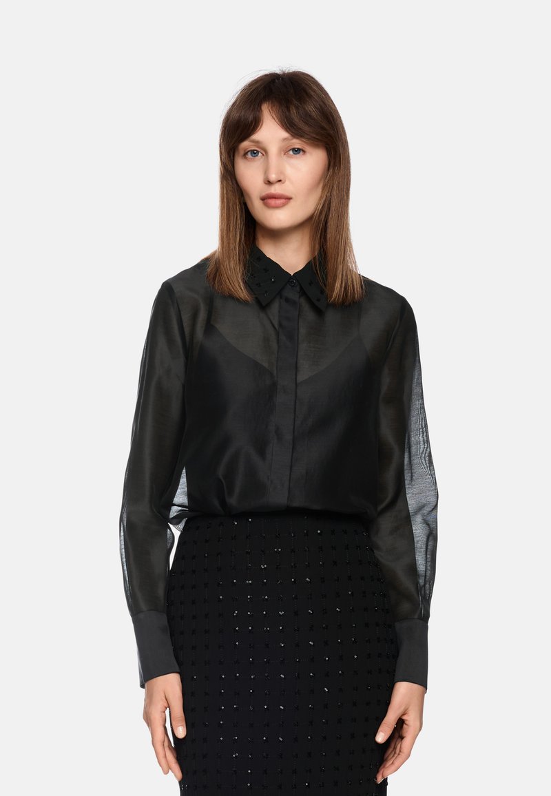 Schwarze, durchsichtige Bluse mit strukturiertem Kragen, detaillierter Knopfleiste und langen Ärmeln. Kombiniert mit einem schwarzen, strukturierten Rock mit Perlenakzenten.