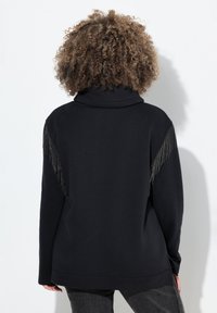 Sudadera negra con cuello alto y detalles metálicos desflecados en los hombros. Tejido suave, corte relajado y mangas largas.