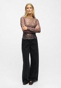 Top à manches longues en tissu brun transparent avec un motif à pois, associé à un pantalon large taille haute noir et des chaussures sombres.