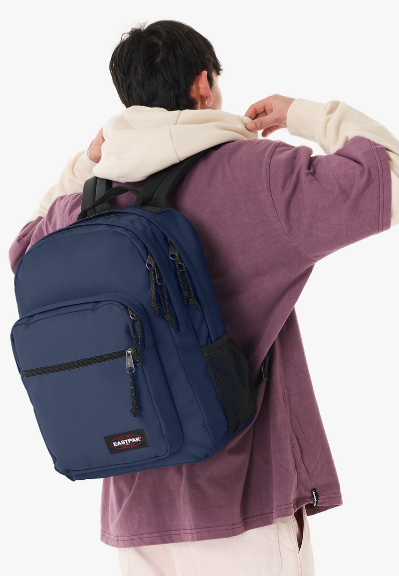 Personne portant une chemise violette et un sweat à capuche beige porte un sac à dos Eastpak bleu marine avec plusieurs compartiments zippés.