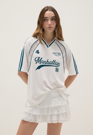 Junge Frau trägt ein weißes, übergroßes Sporttrikot mit dem Schriftzug "Manhattan" und Spitzen-Details, kombiniert mit einem mehrlagigen weißen Spitzrock, und steht vor einem einfarbigen Hintergrund.