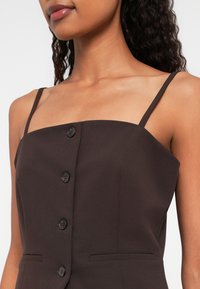 Sieviete, tērpusies tumši brūnā bezpiedurknju topā ar plānām plecu siksnām un pogu detaļām priekšpusē, parādot kaklu un augšējo krūšu daļu.