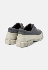 Zapatos de cuero gris con una suela blanca gruesa y estriada, que presentan el logo "Camper" en el talón. Los zapatos se muestran desde la parte de atrás.