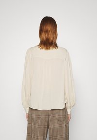 Persona con capelli castani di lunghezza spalle, che indossa una blusa crema larga e pantaloni a quadri marroni, vista da dietro su uno sfondo semplice.