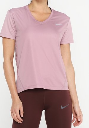 Kobieta w jasno fioletowej koszulce Nike z krótkim rękawem i dekoltem w serek oraz ciemnobrązowych legginsach Nike, stoi z rękami swobodnie opuszczonymi wzdłuż ciała.