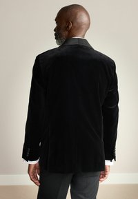 Zwarte fluwelen blazer met een gladde textuur, voorzien van een satijnen kraag, getailleerde pasvorm en drie knopen op de mouwen.