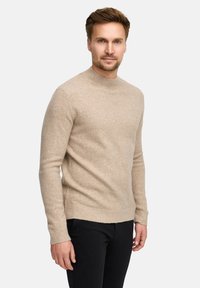 Maglione beige in maglia con scollo rotondo, maniche lunghe e polsini a coste. Texture morbida, design aderente, abbinato a pantaloni neri.