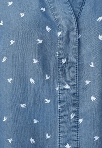 Jeansskjorta med en ljusblå bas som har vita fågelmönster, en v-ringning och synlig sömnad längs knäppslån och sömmarna.