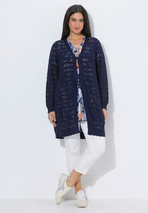 Kvinde iført en marineblå åben strikket cardigan over en blomsterbluse, hvide korte bukser og hvide sneakers, stående mod en hvid baggrund.