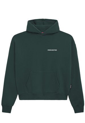 Joven con sudadera con capucha verde oscuro con bolsillo frontal y vaqueros azul claro, de pie frente a un fondo liso gris claro.