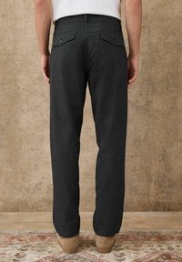 Person wearing black textured trousers with buttoned back pockets and beige suede shoes, standing on a patterned rug against a beige wall.