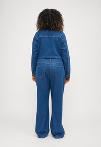 Salopette en denim, bleu clair, avec des manches longues, un pantalon large, des poches arrière et des détails de couture contrastants. Texture lisse.
