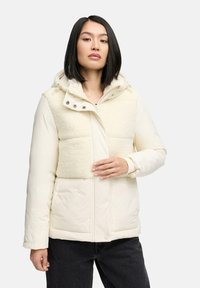 Pufferjack in crème met een getextureerd, fluffy bovenstuk. Voorzien van een capuchon, een ritssluiting aan de voorkant en zijzakken. Materiaal lijkt zacht en geïsoleerd.