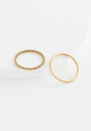 Zwei goldene Ringe auf einer weißen Oberfläche: einer ist verdreht und rund, der andere ein einfacher, glatter Ring, beide mit glänzendem Metallfinish.