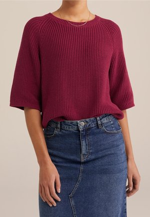 Femme portant un pull en maille bordeaux texturé avec des manches jusqu’aux coudes, associé à une jupe en jean bleue taille haute, debout devant un fond neutre.
