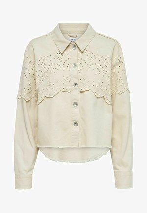 Jasny beżowy krótki top z zapięciem na guziki, kołnierzykiem i koronkowym haftem w kształcie fal na rękawach. Posiada również postrzępione wykończenie u dołu.