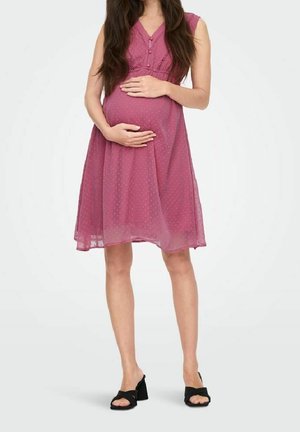 Mujer embarazada con un vestido rosa sin mangas hasta la rodilla con puntos texturizados, sosteniendo su vientre, y usando sandalias negras de tacón con dedos abiertos.