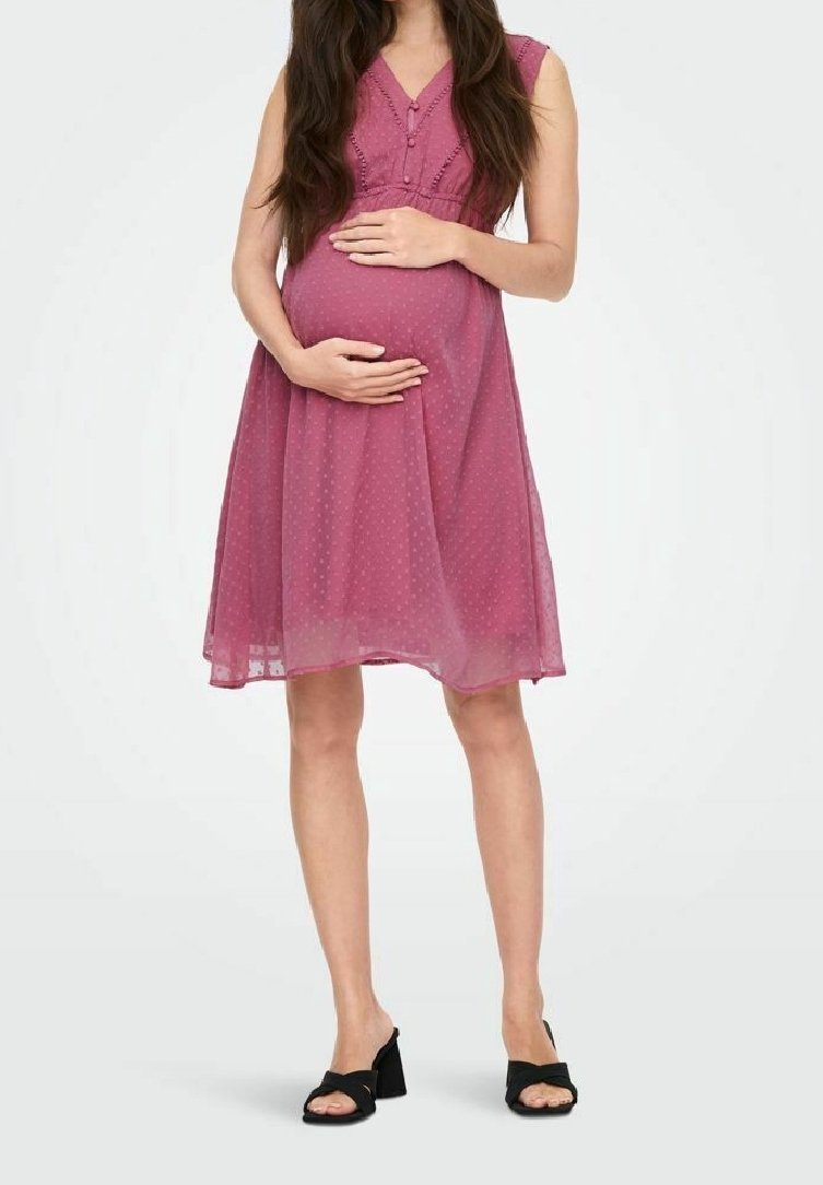 Femme enceinte en robe rose sans manches longueur genou avec des points texturés, tenant son ventre, portant des sandales noires à talons ouvertes.