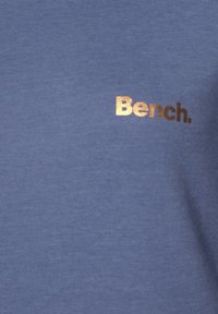 T-shirt in cotone blu con una sottile texture e un logo "Bench." in bronzo metallico sul fronte. Maniche corte, design a girocollo.