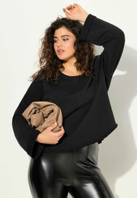 Felpa nera oversize con maniche larghe e arrotondate, abbinata a leggings neri lucidi. Tiene una borsa piegata beige con dettagli a bottoni.