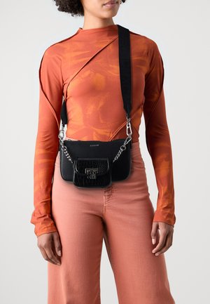 Schwarze Kunstleder-Crossbody-Tasche mit einem silbernen Kettendetail und einem kleineren, strukturierten Beutel; getragen mit einem enganliegenden orangefarbenen Oberteil und hellrosa Hosen.