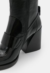 Bottine en cuir noir avec une finition brillante, talon bloc angulaire, fermeture éclair sur le côté et détails de sangle au niveau de la cheville pour un maintien sécurisé.