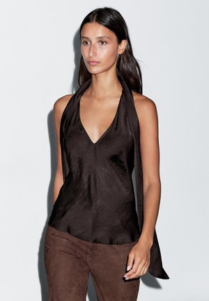 Hnědý halter top s hlubokým výstřihem do "V", vyrobený z texturované látky, se dlouhými vázacími pásky, které sahají dolů po zádech. Kombinovaný s hnědými semišovými kalhotami.