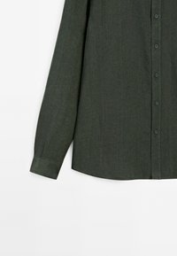 Dark green long-sleeve shirt made of textured fabric, featuring a buttoned front and a curved hemline. Includes buttoned cuffs.