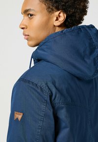 Chubasquero ligero de color navy con capucha, que cuenta con una tela texturizada, costuras visibles y un logo de cuero de Wrangler en el hombro izquierdo.