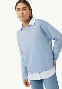 Lichtblauwe sweatshirt met een relaxte pasvorm en geborduurde tekst. Gedragen over een wit overhemd met kraag, gecombineerd met lichte spijkerbroeken.