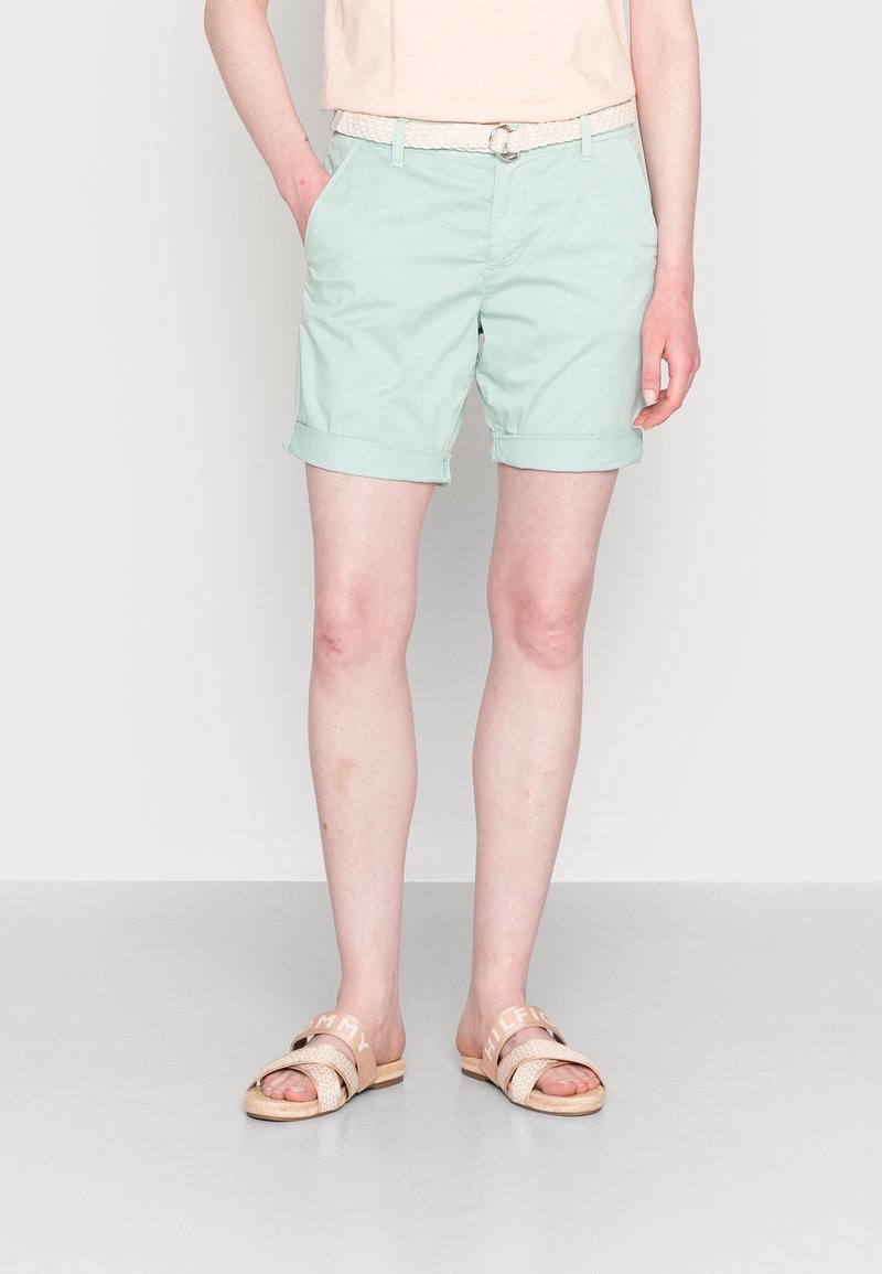 Personne portant un short bleu clair à ourlet retroussé avec une ceinture blanche tressée, un haut rose pâle et des sandales beiges à bouts ouverts, debout les mains dans les poches.