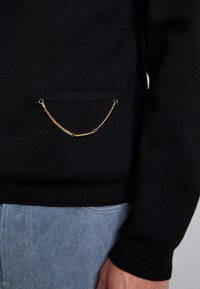 Maglione nero lavorato a maglia con una piccola tasca frontale accentuata da una sottile catena dorata, indossato con jeans in denim azzurro chiaro.