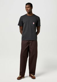 Zwart T-shirt met korte mouwen, voorzien van een borstzak en logo, gecombineerd met losse bruine broek. Bruine sneakers maken de outfit compleet.