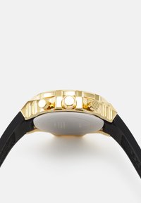 Goldfarbene Uhr mit strukturiertem Gehäuse, drei Funktionsknöpfen an der Seite und einem schwarzen Gummiband. Hat eine glatte Rückseite ohne sichtbare Details.