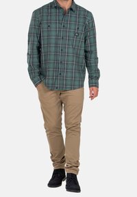 Chemise en flanelle à carreaux verts avec boutons, manches longues, deux poches poitrine, associée à un pantalon beige et des chaussures noires.