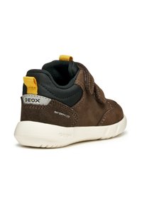 Baskets en suède marron et textile noir avec une semelle en caoutchouc blanc, dotées d'une sangle Velcro et d'une languette jaune. Étiquette "WATERPROOF" sur le côté.