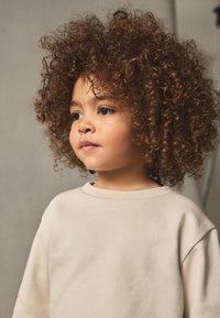 Beige collegepaita, jossa on pyöreä pääntie ja pitkät hihat. Paita on päällä lapsella, jolla on runsaat kiharat hiukset. Tausta on neutraali.
